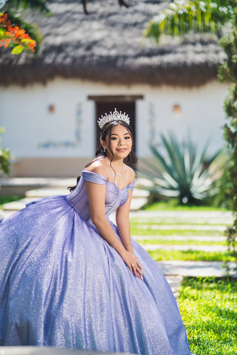 Quinceañera con vestido lila y corona frente a una casa de palma